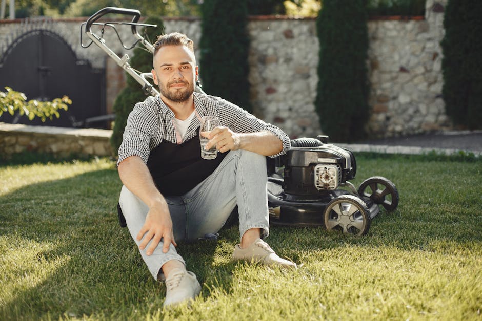 A man with light skin and dark hair, styled with a beard, is sitting barefoot on a well-maintained grassy garden lawn, holding a glass of water and smiling at the camera. He is wearing a casual checke